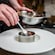 A chef plating food using a metal ring mold and spoon in a professional kitchen